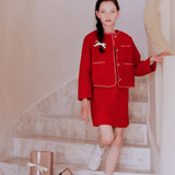 A girl in the KIKI and JOJO Girls Tweed Jacket - Cranberry Red with champagne gold buttons and white shoes stands on a staircase beside several wrapped gift boxes.