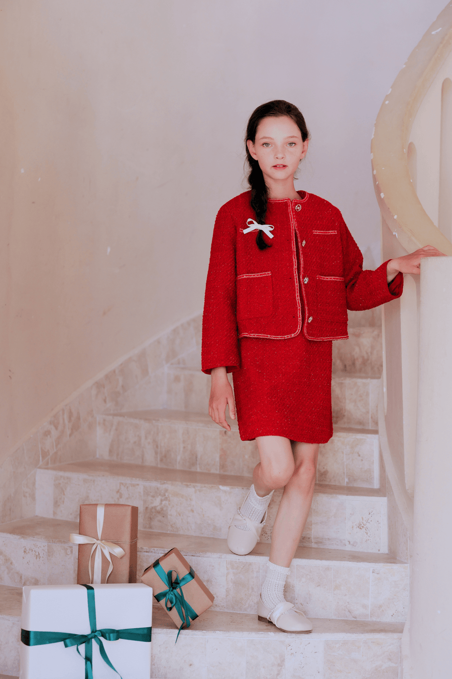A girl in the KIKI and JOJO Girls Tweed Jacket - Cranberry Red with champagne gold buttons and white shoes stands on a staircase beside several wrapped gift boxes.