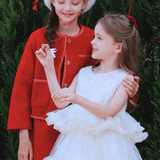 Two young girls smile outdoors before lush greenery; one wears the KIKI and JOJO Girls Tweed Jacket in Cranberry Red with champagne gold buttons, while the other is dressed in a white ruffled dress.