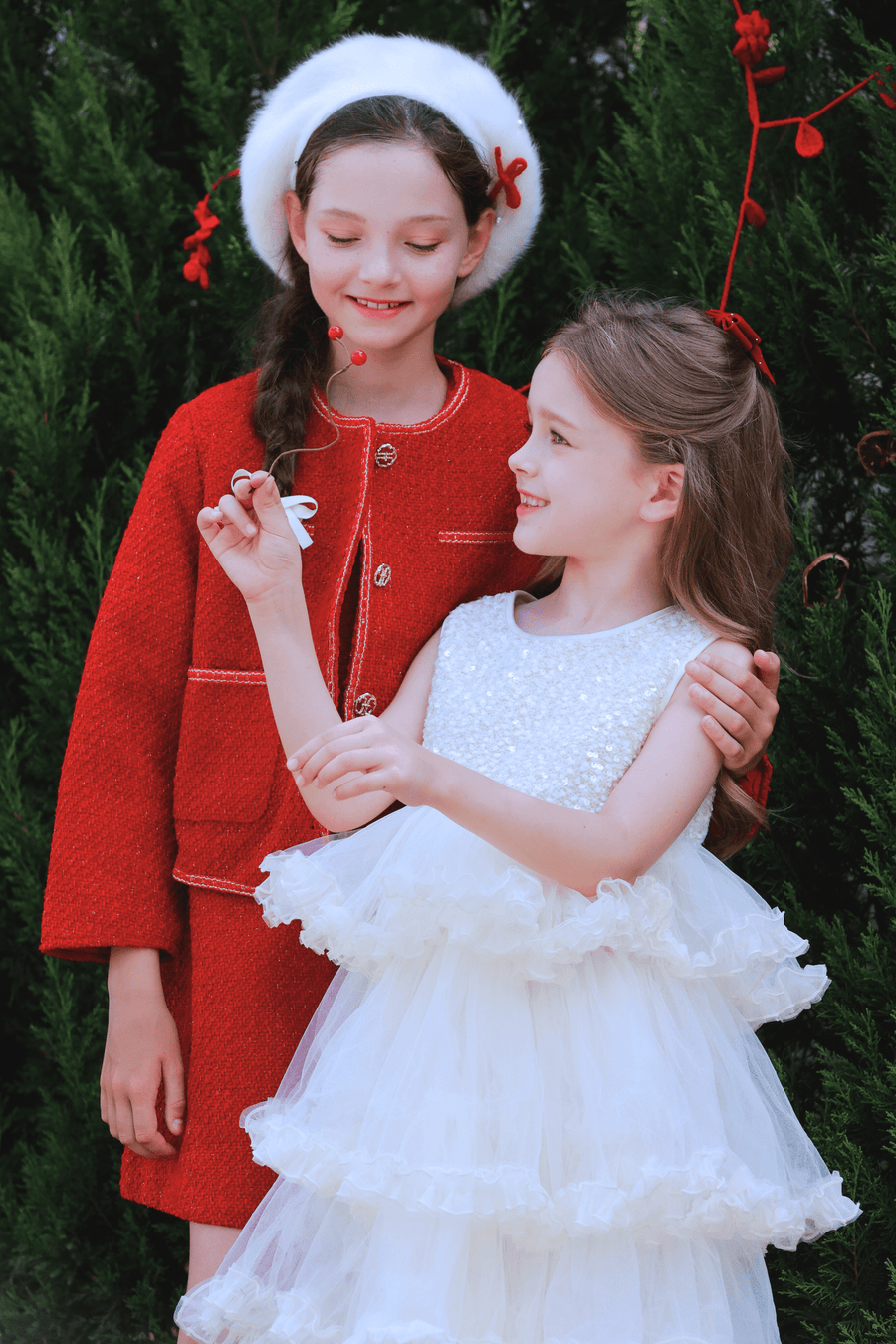 Two young girls smile outdoors before lush greenery; one wears the KIKI and JOJO Girls Tweed Jacket in Cranberry Red with champagne gold buttons, while the other is dressed in a white ruffled dress.