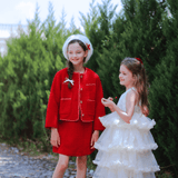 Two girls stand outdoors amid green trees, smiling. One wears the KIKI and JOJO Girls Tweed Jacket in Cranberry Red with a white beret; the other wears a white ruffled dress.
