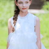 A young girl with a braided hairstyle wears the KIKI and JOJO Girls Silk Satin Jeweled & Padded Headband in champagne, paired with a sleeveless, ruffled white dress while standing outdoors on a sunny day.