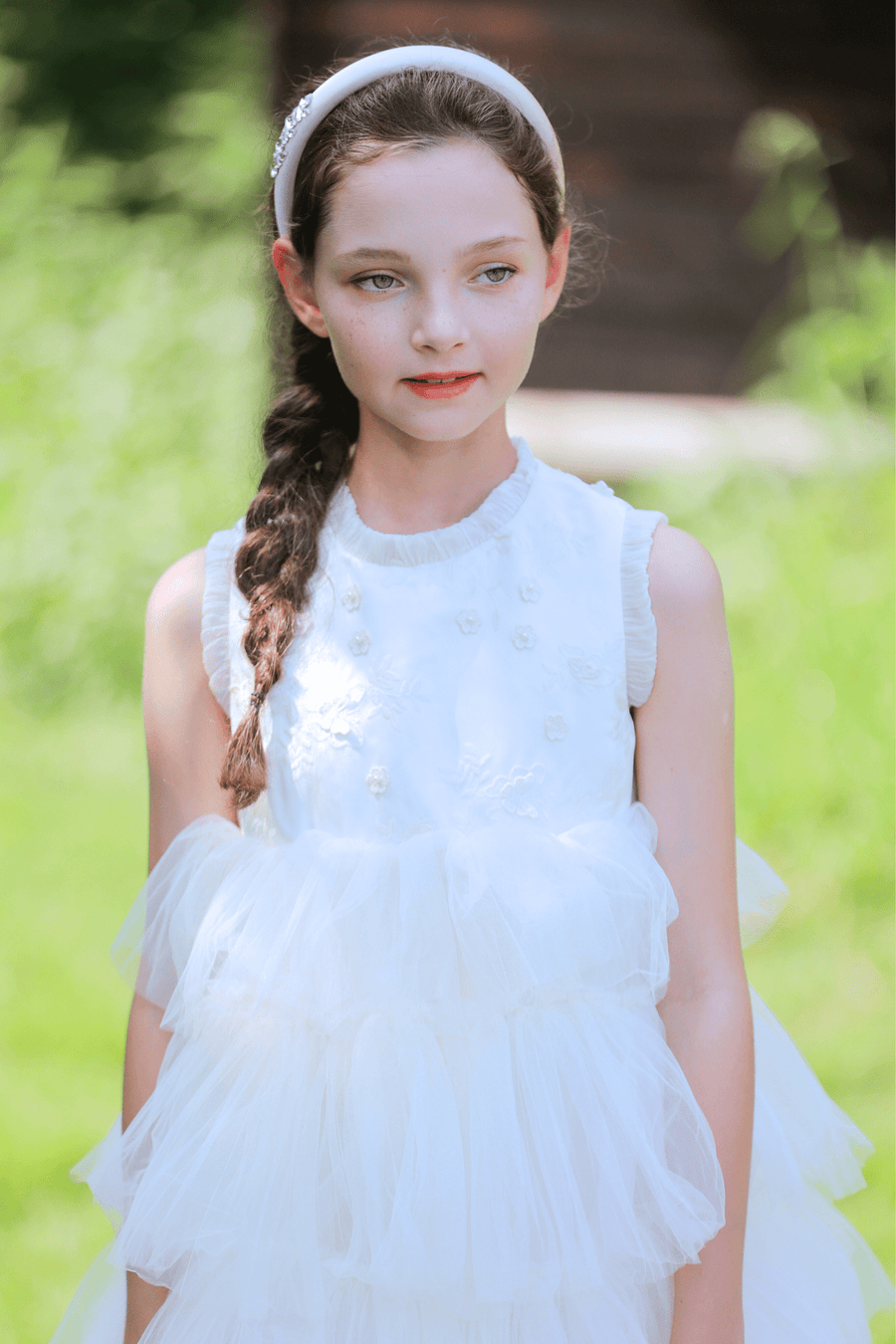 A young girl with a braided hairstyle wears the KIKI and JOJO Girls Silk Satin Jeweled & Padded Headband in champagne, paired with a sleeveless, ruffled white dress while standing outdoors on a sunny day.