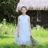 A young girl stands on grass by a thatched-roof building, wearing the KIKI and JOJO Girls Silk Satin Jeweled & Padded Headband in Champagne with a white ruffled dress and holding dried flowers amid lush greenery.