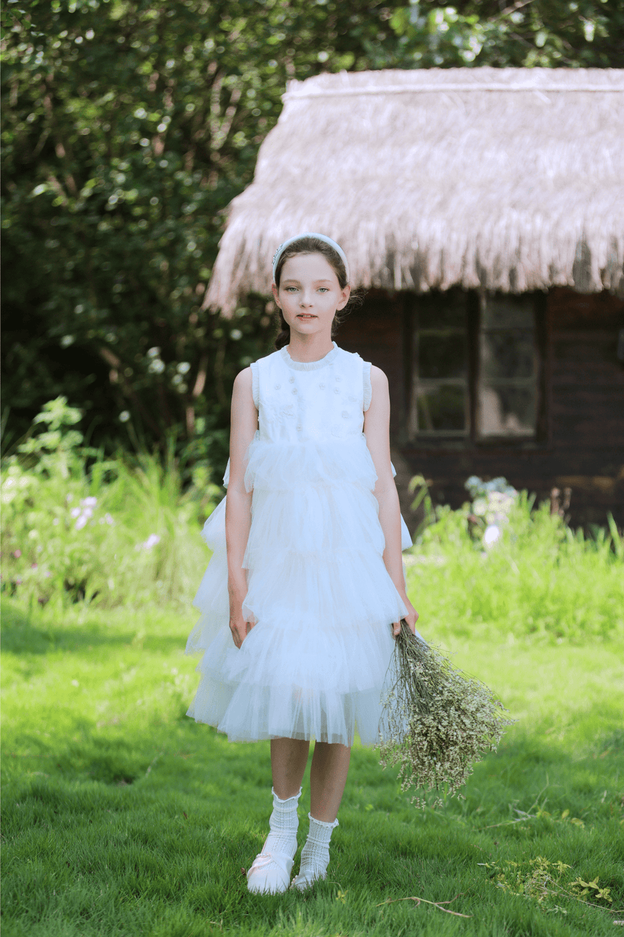 A young girl stands on grass by a thatched-roof building, wearing the KIKI and JOJO Girls Silk Satin Jeweled & Padded Headband in Champagne with a white ruffled dress and holding dried flowers amid lush greenery.
