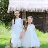 Two girls in white dresses walk on grass, one wearing the KIKI and JOJO Girls Silk Satin Jeweled & Padded Headband in Champagne, in front of a thatched-roof wooden building with lush greenery behind.