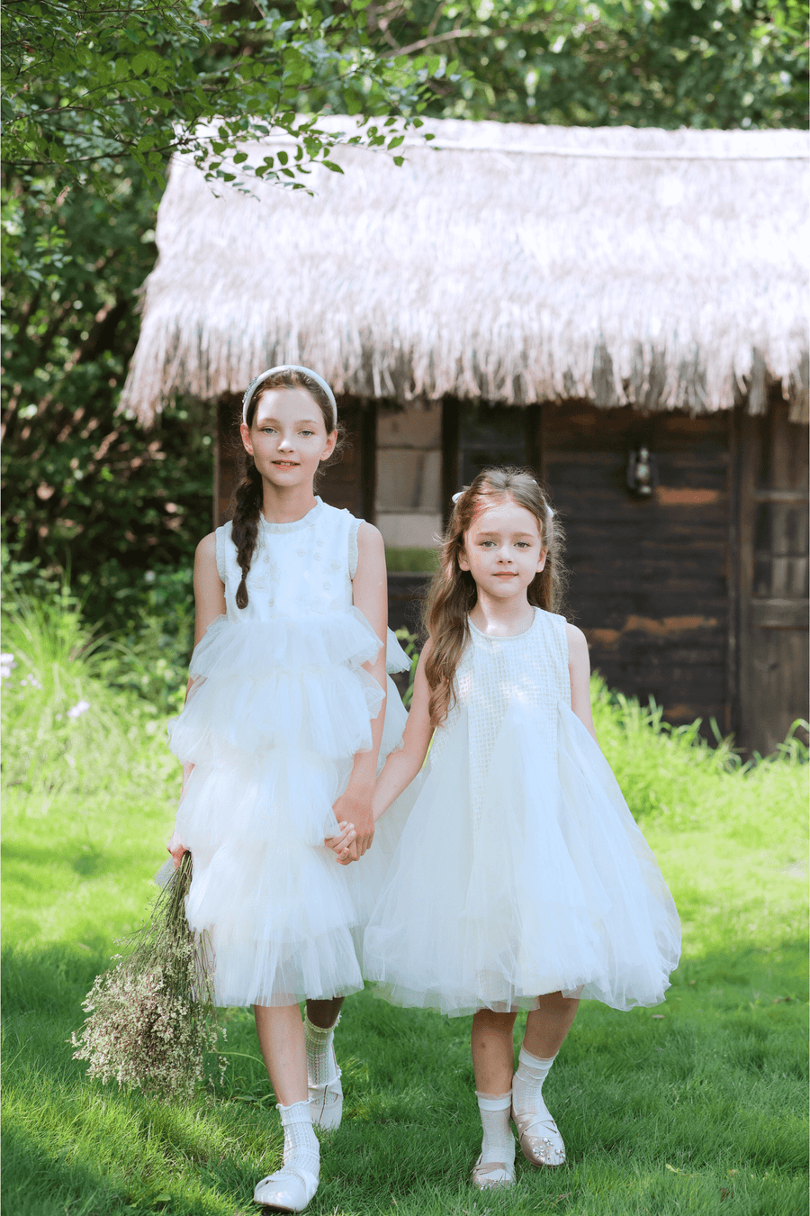 Two girls in white dresses walk on grass, one wearing the KIKI and JOJO Girls Silk Satin Jeweled & Padded Headband in Champagne, in front of a thatched-roof wooden building with lush greenery behind.