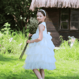 A young girl in a white dress wears the KIKI and JOJO Girls Silk Satin Jeweled & Padded Headband in Champagne, holding a bouquet of flowers while standing on grass before a thatched-roof building, ready for her special occasion.