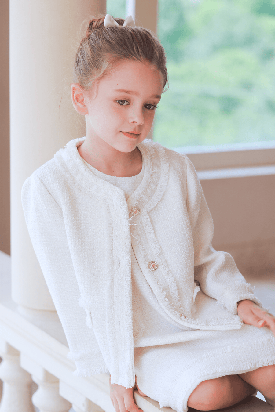 A young girl with light brown hair tied up in KIKI and JOJO Girls Silk Satin Ribbon Bow Hair Clips (2 Pack) in Ivory, wearing a white dress and jacket, sits indoors on a stone ledge with greenery visible through the window.