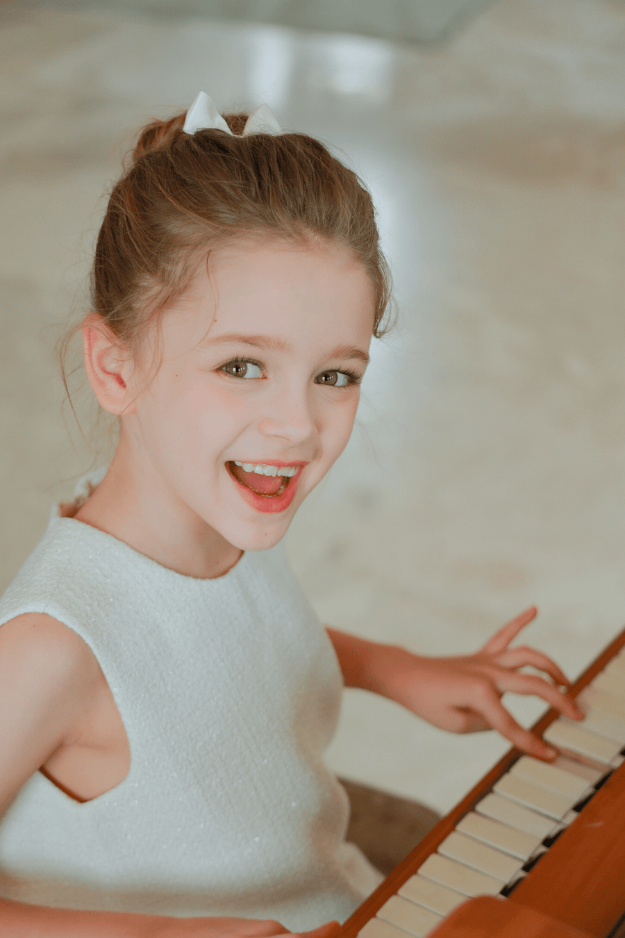 A young girl with light brown hair in a white dress smiles at the piano, her look finished with KIKI and JOJO’s Girls Silk Satin Ribbon Bow Hair Clips (2 Pack) in Ivory—a charming accessory for girls.