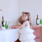 A young girl in a KIKI and JOJO Girls Sparkle Sleeveless Tulle Dress in Champagne stands by a wooden counter adorned with bottles and glassware.