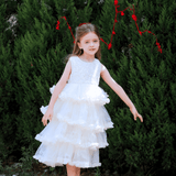 A young girl wears the KIKI and JOJO Girls Sparkle Sleeveless Tulle Dress in Champagne, standing outdoors on a gravel path before tall green bushes, with red string decorations in the background.
