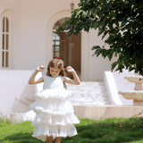 A young girl wears the KIKI and JOJO Girls Sparkle Sleeveless Tulle Dress in champagne as she stands on green grass near a tree, with a white building and fountain visible in the background.