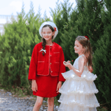 Two young girls stand outdoors; one wears a red outfit with a white beret, the other is dressed in the KIKI and JOJO Girls Sparkle Sleeveless Tulle Dress in Champagne. Green bushes and blue sky provide a charming backdrop.