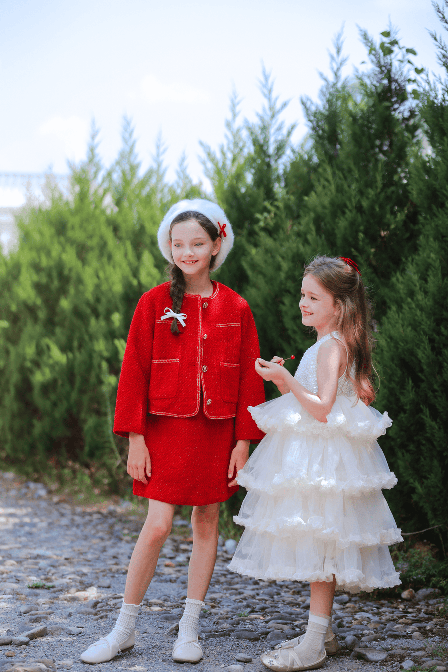 Two young girls stand outdoors; one wears a red outfit with a white beret, the other is dressed in the KIKI and JOJO Girls Sparkle Sleeveless Tulle Dress in Champagne. Green bushes and blue sky provide a charming backdrop.