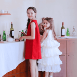 Two young girls stand by a table with bottles and glasses. One wears the KIKI and JOJO Girls Sparkle Sleeveless Tulle Dress in Champagne, while the other is in a white flower girl dress; both look toward the camera.