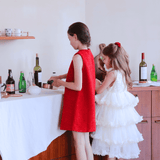 Two young girls in KIKI and JOJO's Girls Sparkle Sleeveless Tulle Dress - Champagne stand at a table with a white cloth, preparing food or drinks among bottles and utensils in a cozy home setting.