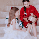 Two young girls in KIKI and JOJO Girls Sparkle Sleeveless Tulle Dresses in Champagne sit on marble stairs; one holds a wrapped gift as the other looks on. Stacked presents with green ribbons are behind them.