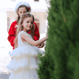 Two young girls stand outside—a girl in the KIKI and JOJO Sparkle Sleeveless Tulle Dress in Champagne touches a bush, while another girl in a red coat and white beret stands behind her. A white building is visible in the background.