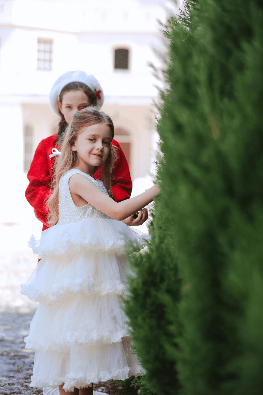 Two young girls stand outside—a girl in the KIKI and JOJO Sparkle Sleeveless Tulle Dress in Champagne touches a bush, while another girl in a red coat and white beret stands behind her. A white building is visible in the background.