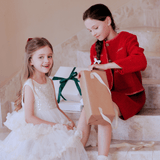 Two young girls sit on marble stairs with gift boxes; one smiles at the camera in a KIKI and JOJO Girls Sparkle Sleeveless Tulle Dress - Champagne, while the other, in a red outfit, opens a brown paper bag.