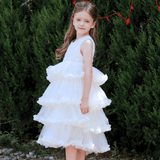 A young girl wears the KIKI and JOJO Girls Sparkle Sleeveless Tulle Dress in Champagne, standing on a gravel path before green bushes and looking at the camera.