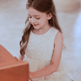 A young girl with long brown hair and a red bow, wearing the KIKI and JOJO Girls Sparkle Sleeveless Tulle Dress in Champagne, sits at a piano and smiles while playing.