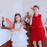 Indoors, one girl in a KIKI and JOJO Girls Sparkle Sleeveless Tulle Dress - Champagne holds bread, while another in a red dress holds a sausage on a fork; bottles and food are on the wooden counter behind them.