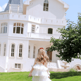 A girl wearing the KIKI and JOJO Girls Sparkle Sleeveless Tulle Dress in Champagne walks across green grass toward a white house with arched windows and a pointed roof on a sunny day.