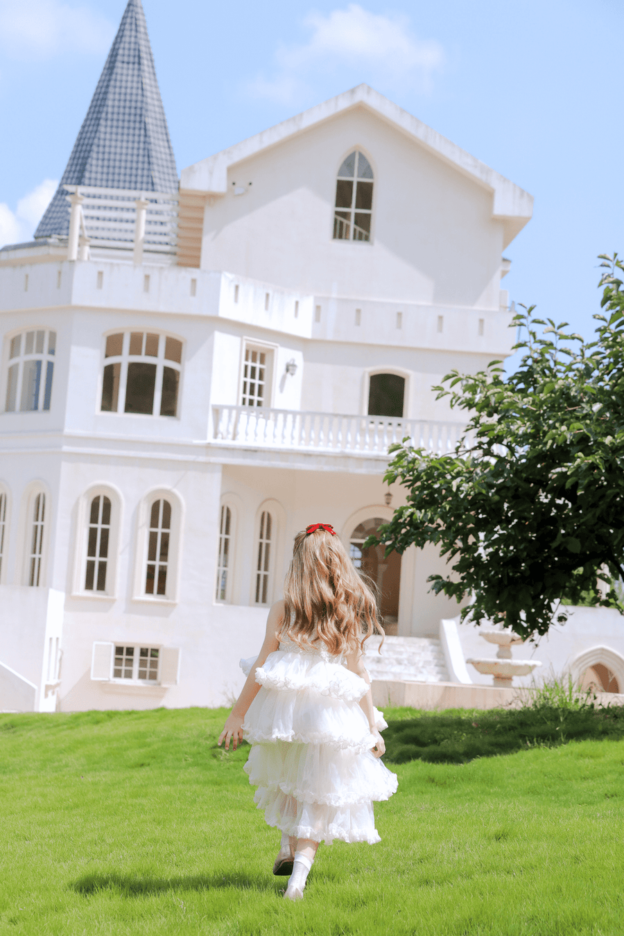 A girl wearing the KIKI and JOJO Girls Sparkle Sleeveless Tulle Dress in Champagne walks across green grass toward a white house with arched windows and a pointed roof on a sunny day.