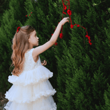 A young girl in a KIKI and JOJO Girls Sparkle Sleeveless Tulle Dress - Champagne hangs red roses on a tall green bush outdoors, her sparkling gown bringing charm to the scene.