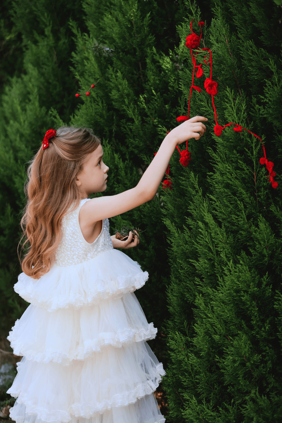 Wearing the KIKI and JOJO Girls Sparkle Sleeveless Tulle Dress in Champagne, a young girl hangs red yarn decorations on a green bush, her dress fluttering as she works.