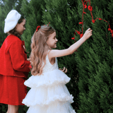 Two young girls, one in a KIKI and JOJO Girls Sparkle Sleeveless Tulle Dress - Champagne and the other in a red coat and beret, decorate an outdoor evergreen bush with red string and ornaments.