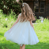 A young girl with long hair, seen from behind, walks on grass toward a rustic thatched-roof house wearing the KIKI and JOJO Girls Textured Velvet Sleeveless Embroidered Dress in Ivory.