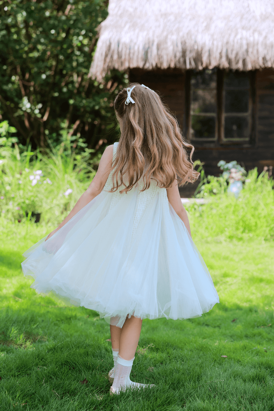 A young girl with long hair, seen from behind, walks on grass toward a rustic thatched-roof house wearing the KIKI and JOJO Girls Textured Velvet Sleeveless Embroidered Dress in Ivory.