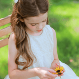 A young girl with long brown hair and white bows wears the KIKI and JOJO Girls Textured Velvet Sleeveless Embroidered Dress in Ivory while sitting on a wooden bench outdoors, looking down and holding a yellow sunflower.