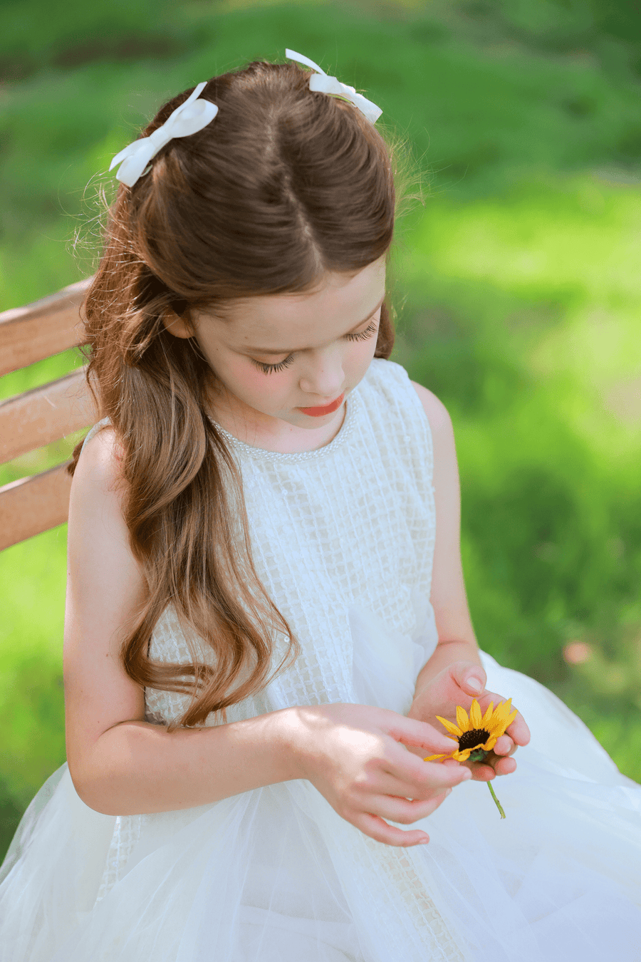 A young girl with long brown hair and white bows wears the KIKI and JOJO Girls Textured Velvet Sleeveless Embroidered Dress in Ivory while sitting on a wooden bench outdoors, looking down and holding a yellow sunflower.