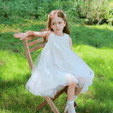 A young girl wears the KIKI and JOJO Girls Textured Velvet Sleeveless Embroidered Dress in ivory as she sits on a wooden chair outdoors on green grass, looking to the side with one arm resting on the chair back.