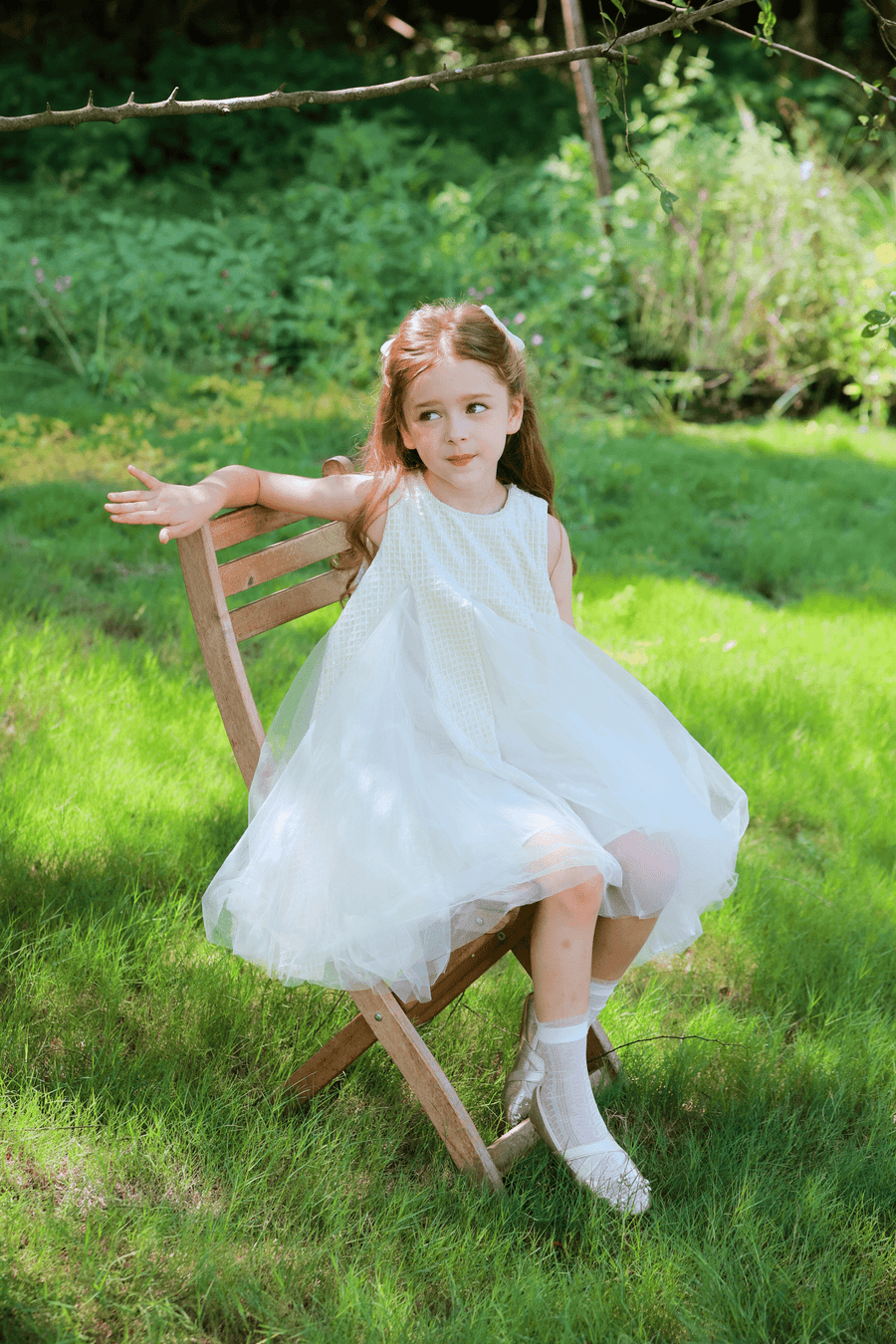 A young girl wears the KIKI and JOJO Girls Textured Velvet Sleeveless Embroidered Dress in ivory as she sits on a wooden chair outdoors on green grass, looking to the side with one arm resting on the chair back.