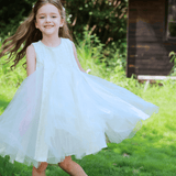 A young girl in a KIKI and JOJO Girls Textured Velvet Sleeveless Embroidered Dress in ivory spins on green grass near a wooden hut, smiling with trees in the background.