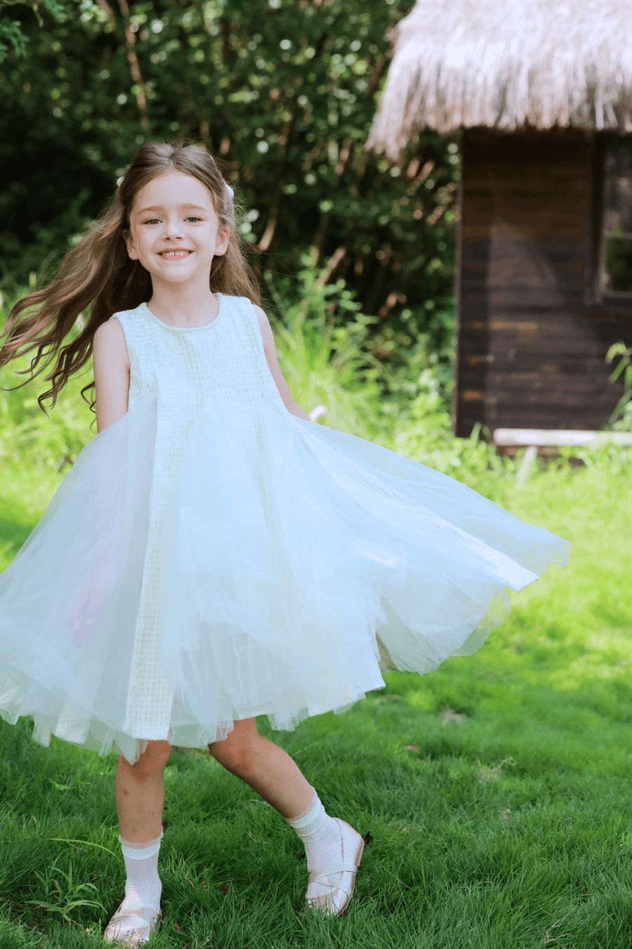 A young girl in a KIKI and JOJO Girls Textured Velvet Sleeveless Embroidered Dress in ivory spins on green grass near a wooden hut, smiling with trees in the background.