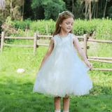 A young girl wears the KIKI and JOJO Girls Textured Velvet Sleeveless Embroidered Dress in Ivory, standing on grass, holding her skirt and smiling with a wooden fence and greenery in the background.
