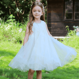 A young girl in the KIKI and JOJO Girls Textured Velvet Sleeveless Embroidered Dress - Ivory stands on grass, holding out her skirt, with a wooden house and lush greenery behind her.