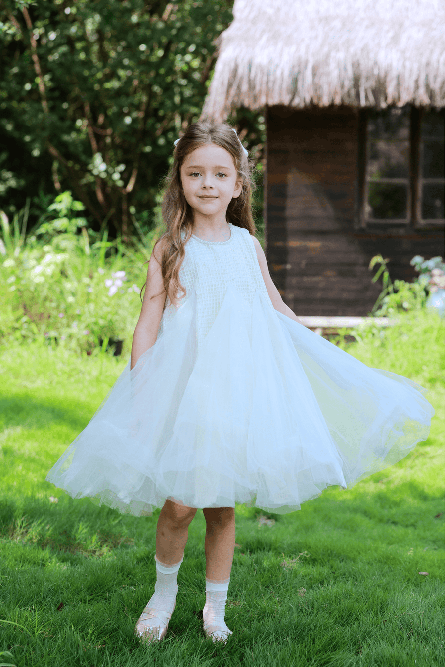 A young girl in the KIKI and JOJO Girls Textured Velvet Sleeveless Embroidered Dress - Ivory stands on grass, holding out her skirt, with a wooden house and lush greenery behind her.