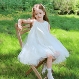 A girl wearing the KIKI and JOJO Girls Textured Velvet Sleeveless Embroidered Dress in ivory sits on a wooden chair outdoors, surrounded by lush greenery.