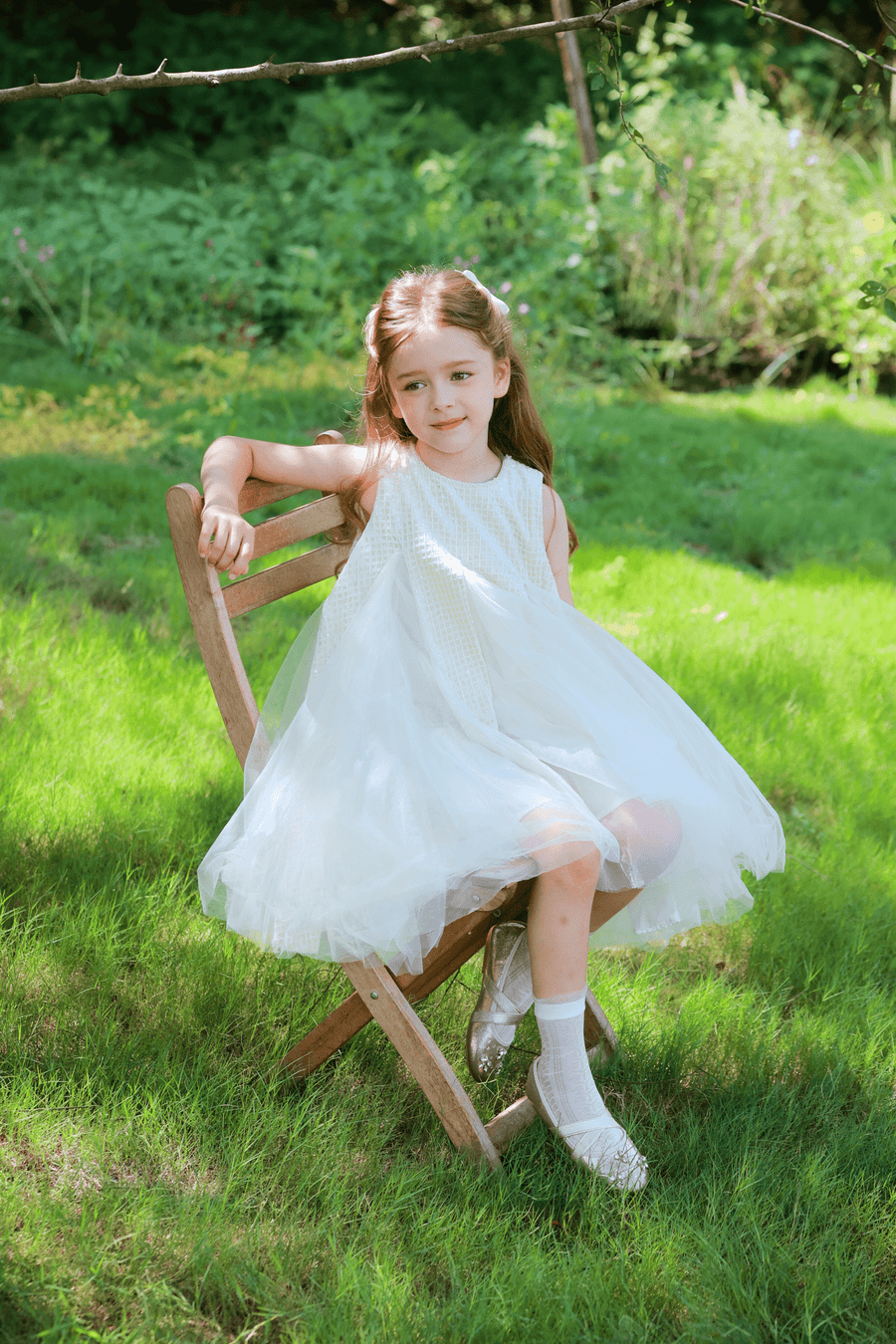 A girl wearing the KIKI and JOJO Girls Textured Velvet Sleeveless Embroidered Dress in ivory sits on a wooden chair outdoors, surrounded by lush greenery.