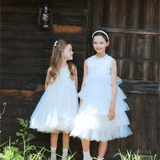Two young girls wear KIKI and JOJO Girls Textured Velvet Sleeveless Embroidered Dress in Ivory, standing side by side before a rustic wooden building with a thatched roof and lantern, surrounded by green grass.