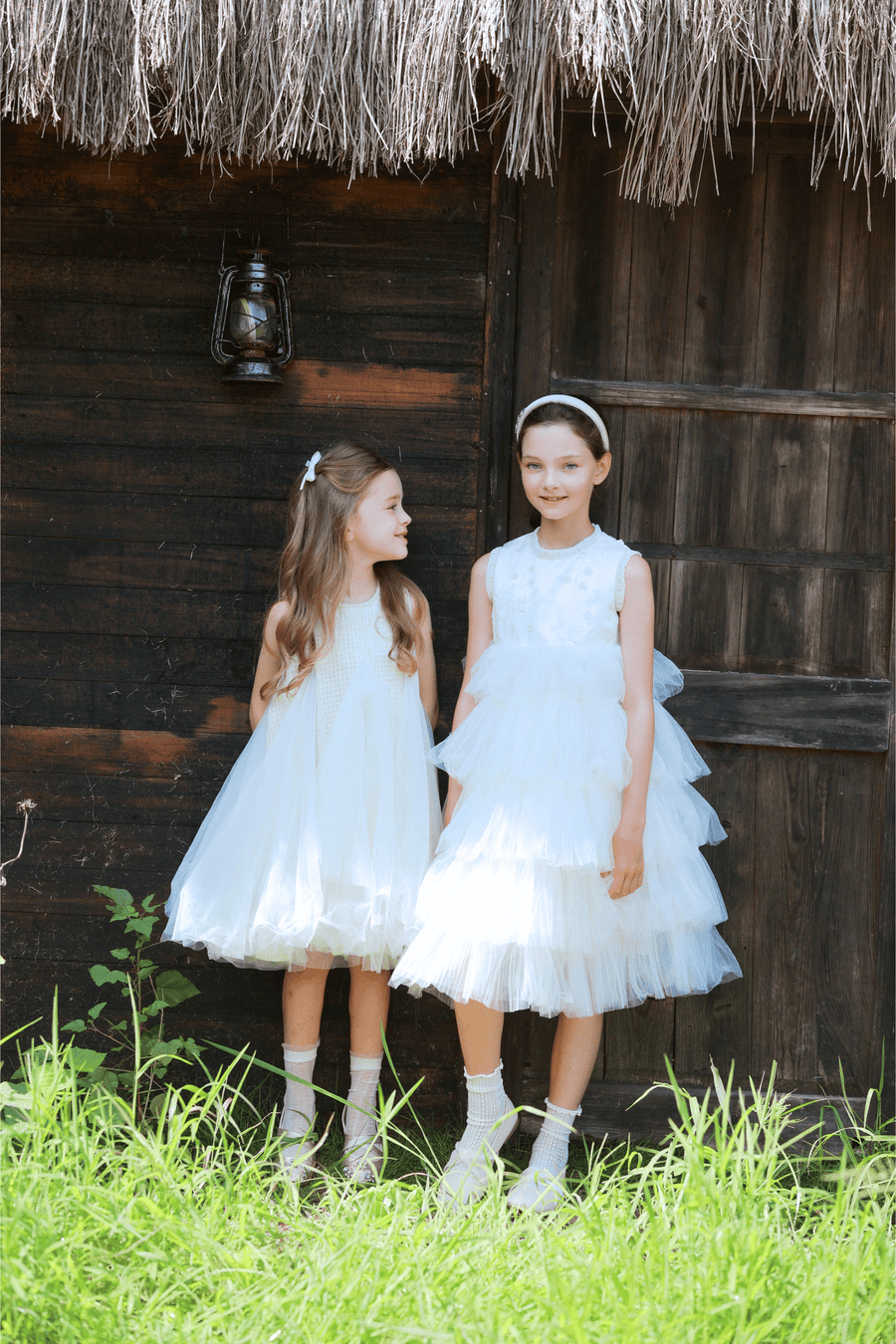 Two young girls wear KIKI and JOJO Girls Textured Velvet Sleeveless Embroidered Dress in Ivory, standing side by side before a rustic wooden building with a thatched roof and lantern, surrounded by green grass.