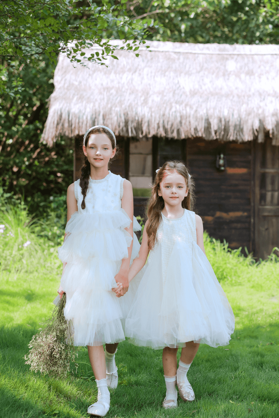 Two young girls in KIKI and JOJO Girls Textured Velvet Sleeveless Embroidered Dress in Ivory hold hands while walking on grass near a wooden cottage with a thatched roof, surrounded by greenery.