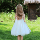 A young girl wears the KIKI and JOJO Girls Textured Velvet Sleeveless Embroidered Dress in Ivory, standing on green grass with a wooden hut and dense foliage in the background, facing away from the camera.